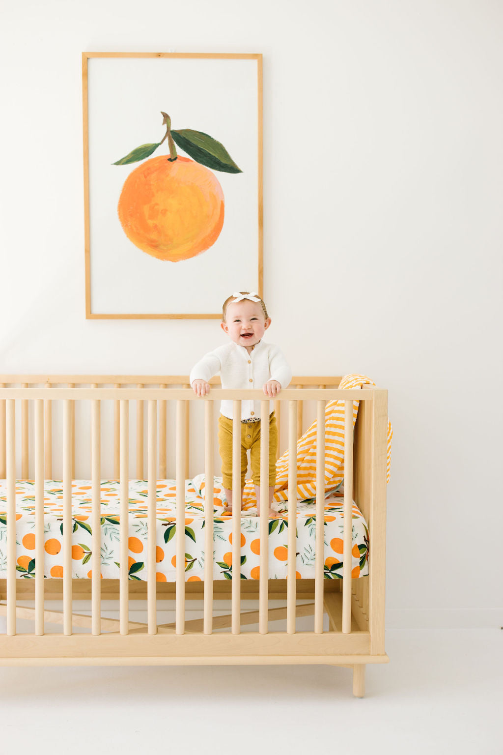Child standing in a crib with orange-themed bedding and a framed picture of an orange on the wall.